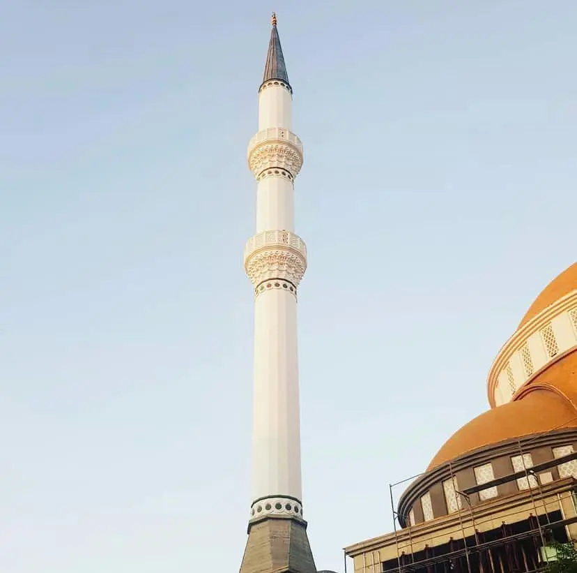 Çift şerefeli minare — mukarnas bilezikleri, koyu külah ve altın alem; sağda inşa hâlindeki kubbe