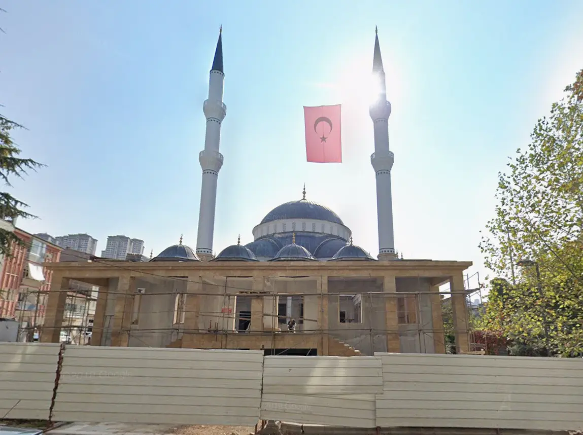 İnşaat aşaması — yükselen iki minare, taş kaplı gövde ve kurşun tonlu kubbeler, aralarında Türk bayrağı